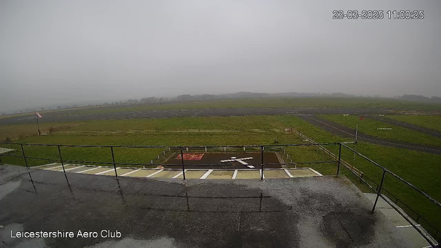 A view from a webcam at Leicestershire Aero Club showing a foggy landscape. In the foreground, there is a small helipad marked with a white cross and bordered by wooden fencing. The background features grassy fields extending into the distance, with a narrow runway visible. The sky appears overcast and gray, creating a misty atmosphere. The image is timestamped with the date and time in the top right corner.