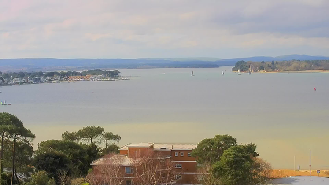 A panoramic view of a harbor with boats sailing on calm waters. In the foreground, there are trees and a building with a reddish facade. The background features a shoreline with various sailboats and distant hills under a cloudy sky. The water transitions from lighter shades near the shore to darker hues further out.