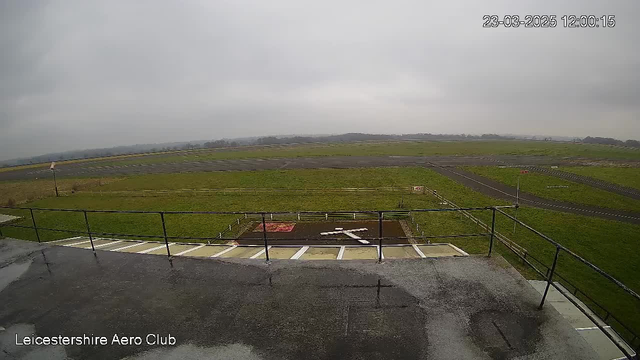 A gray and overcast sky is visible above a large, grassy aerodrome. In the foreground, there is a flat rooftop area with a safety railing. Below the rooftop, a landing area is marked with a white cross and a red square on brown earth. The aerodrome has a paved runway visible in the background, bordered by green grass and wooden fencing. The scene appears calm and quiet with no activity evident.