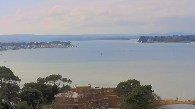 A scenic view of a calm body of water, with several small boats and sailboats visible. In the background, a shoreline lined with trees, buildings, and a stretch of beach. The sky is partly cloudy with soft, pastel hues. The foreground features a few trees and a red brick building. The overall scene conveys a peaceful and pastoral atmosphere.