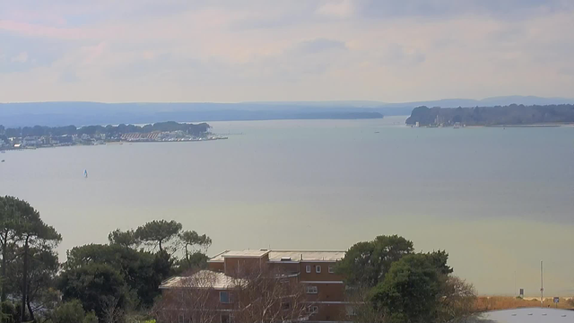 A tranquil view of a body of water, with gentle waves reflecting soft sunlight. In the foreground, there are some trees and a low building with a flat roof. In the distance, the shoreline features a mix of marinas, boats, and houses. The horizon is lined with rolling hills under a slightly cloudy sky, creating a peaceful, serene atmosphere.