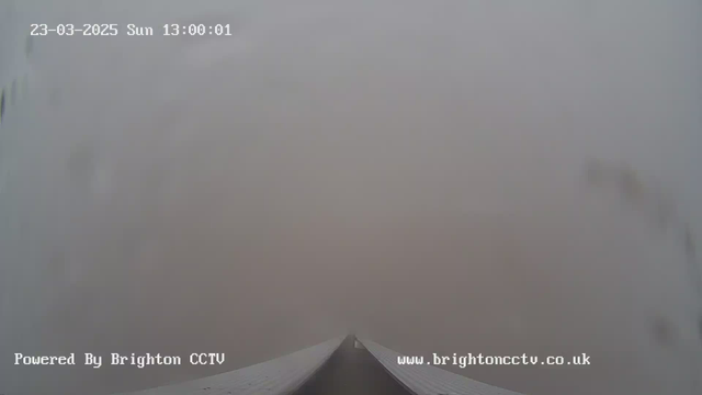 A foggy scene captured by a webcam, showing a long, flat surface extending towards the horizon. The visibility is very low, with gray tones dominating the image. The date and time read "23-03-2025, Sunday, 13:00:01," with a watermark stating "Powered By Brighton CCTV" at the bottom.