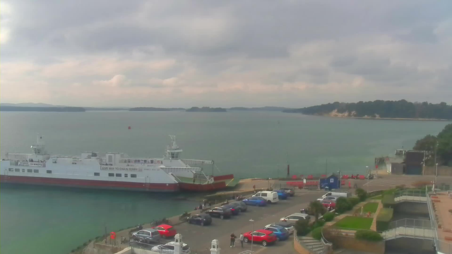 A view of a harbor with a large white ferry docked at the left side. In front of the ferry, there is a parking area filled with various cars, predominantly in shades of red, blue, and gray. The water is calm and stretches into the distance, where a small buoy is visible. The sky is partially cloudy with light gray clouds, and a hilly coastline can be seen on the far side of the water. The scene is tranquil, with a few people milling around near the ferry.