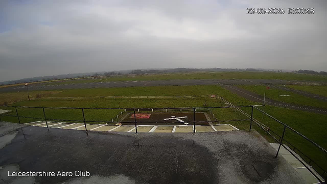A view from a webcam overlooking an airfield. In the foreground, there is a flat rooftop with a railings. Below, the grass-covered field features a large white "X" marking in the center, surrounded by a dirt patch. The area is bordered by a wooden fence, and there are several paved runways visible in the background, stretching towards a gray, cloudy sky. The image is timestamped March 23, 2025, at 1:00:48 PM.