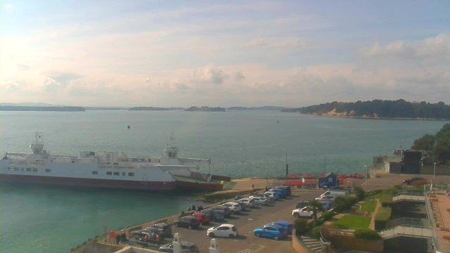 A view of a tranquil harbor with a ferry docked on the left side. The water is calm and reflects the sky, which is mostly clear with some clouds. In the foreground, there is a parking lot filled with various cars, including red, blue, and white vehicles. Green grass and a landscaped area with plants are visible to the right, and a small building is located near the water's edge. In the background, distant trees and hills can be seen across the water, contributing to the serene atmosphere of the scene.