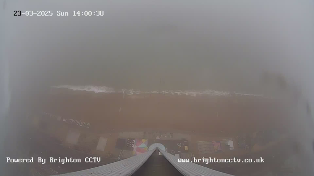 A foggy coastal view captured from a high vantage point. The sandy beach is mostly obscured by mist, with only faint outlines of waves breaking at the shore. Various beach amenities, such as colorful umbrellas and seating areas, are visible along the foreground. The date and time display is at the top of the image, and there is a watermark indicating the location is powered by Brighton CCTV.