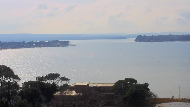 A serene view of a calm body of water extending into the horizon, reflecting sunlight and displaying a shimmering surface. In the foreground, there are some trees and a low building with a flat roof. The distant shoreline features trees and buildings, gradually fading into a misty landscape with rolling hills and a cloudy sky.
