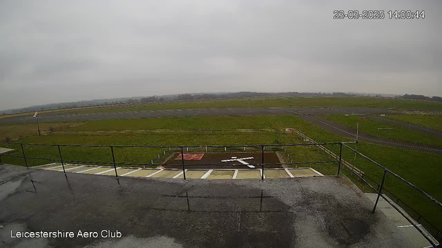 A view from a high vantage point at Leicestershire Aero Club shows a vast expanse of grassy field under a cloudy sky. In the foreground, there is a railing, and below, a runway area features a prominent white cross shape and a red patch of soil. The landscape extends into the distance, bordered by greenery and a few structures along the edges. The overall scene is grey and overcast, suggesting a cloudy day.