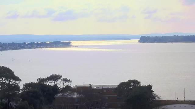 A scenic view of a calm body of water extending towards distant hills under a cloudy sky. The sunlight reflects off the water, creating a shimmering surface. In the foreground, there are trees and a low building, while the shoreline in the background features some boats and patches of land. The overall atmosphere is serene and tranquil.