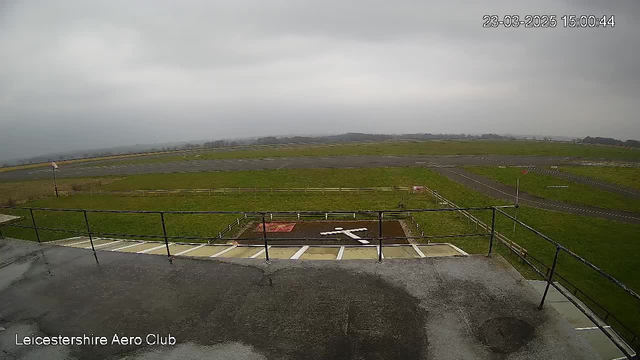 A cloudy sky with grey overcast covers the scene. In the foreground, there is a view from a raised platform looking down at an airfield. The airfield is primarily a green grass area with a dark asphalt runway stretching horizontally across the middle. A large white cross is marked on the grass, indicating a landing spot. There are some low wooden fences outlining parts of the area. A red sign can be seen near the right side of the image. The environment appears calm with no visible aircraft or activity.