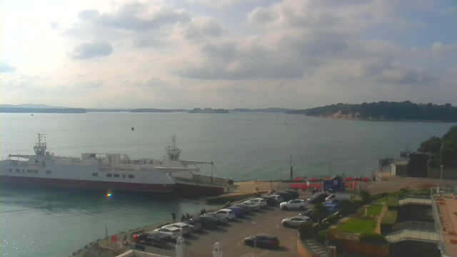A view of a harbor with a ferry docked on the left and several parked cars in the foreground. The water is calm and reflects the sky, which is mostly cloudy with some light appearing through. On the right, there are some buildings and greenery, while distant hills can be seen across the water. A few small boats are visible on the water.
