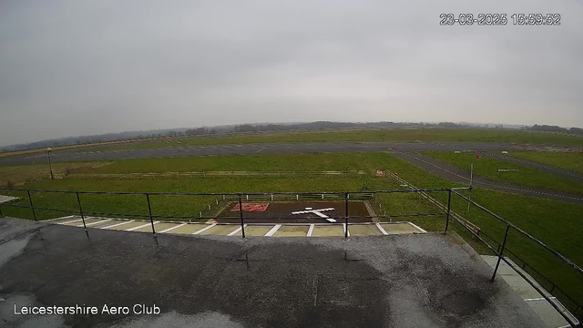 A view from a webcam at the Leicestershire Aero Club, showing an expansive grass field and a runway under a cloudy sky. In the foreground, there is a fenced area with a white cross marking on the ground, surrounded by a concrete pathway. The background features a longer runway with a section of road bordered by a fence.