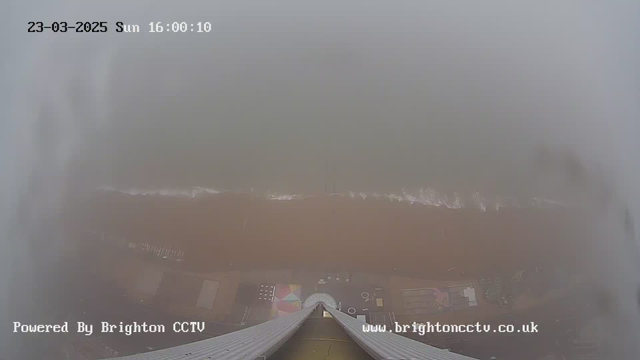 A view from a high angle overlooking a beach on a foggy day. The shoreline is partially visible with gentle waves lapping at the sandy beach, which has a brownish tint. There are various shapes and structures on the beach below, but details are obscured by the fog. The date and time stamp in the top left corner indicates March 23, 2025, at 16:00:10. The bottom of the image features the text "Powered By Brighton CCTV" and a website address, both displayed in white font.