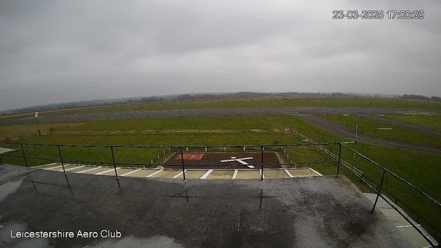A view from a webcam at the Leicestershire Aero Club, showing a cloudy sky and a general aviation airfield. In the foreground, there is a fenced area with some markings on the ground, including a large white cross. The grassy landscape stretches out into the distance, where the airfield is visible with a runway and taxiways. The scene appears overcast, and the time displayed is 5:00 PM.