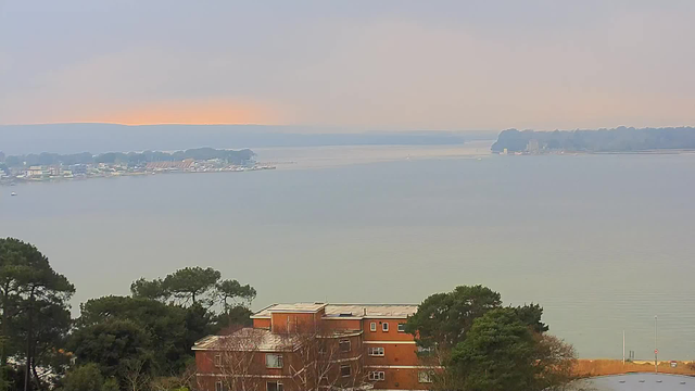 A view from a webcam overlooking a calm body of water at dawn. In the foreground, there are trees and a large building with a reddish brick façade. The background features a shoreline dotted with small houses and boats, and a faint line of orange glow on the horizon signaling the sunrise. The water appears smooth with soft ripples, and there is a pale, cloudy sky above.