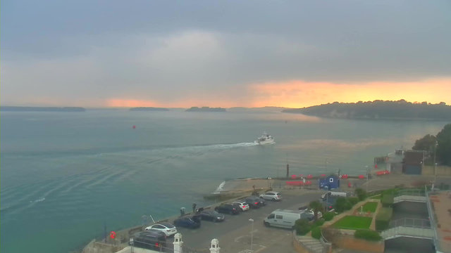 A coastal scene showing a calm ocean under a cloudy sky. A small white boat is leaving a wake as it moves towards the horizon. The water reflects shades of blue and green, while a warm orange glow from the setting sun is visible in the distance. In the foreground, there is a harbor area with parked cars and a path leading to a shoreline. Some people are standing near the water's edge, and several red buoys can be seen floating in the water.