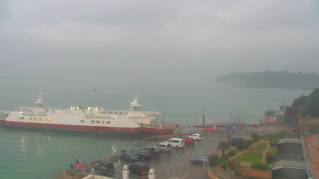 A grey and foggy scene at a harbor. A large white ferry with red accents is docked at the left side of the image, partially obscured by mist. The water is calm but appears subdued in color. A few vehicles are parked along the waterfront, and some people can be seen near the water's edge. In the background, a small green hill is blurred by the fog, and the overall atmosphere is overcast and damp.