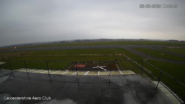 A view from a webcam at Leicestershire Aero Club shows a runway and grassy field under a cloudy sky. There is a cross-shaped landing area marked in white on the ground, and a fence running along part of the field. The image is taken from an elevated position, providing a broad perspective of the landscape.