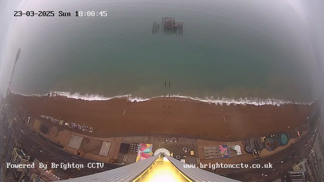 A view from a high vantage point looking down at a beach with golden sand, where waves gently lap at the shore. There are a few structures on the beach and some colorful patterns on the sand, suggesting amusement attractions. To the left, a wooden pier extends into the sea, partially submerged. The sky appears overcast, creating a muted atmosphere. The image has a timestamp in the upper left corner indicating the date and time.