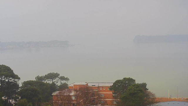 A foggy view of a calm body of water with distant land visible on the horizon. In the foreground, several trees and a brick building are partially visible. The overall scene has a muted color palette due to the haze, creating a tranquil and subdued atmosphere.