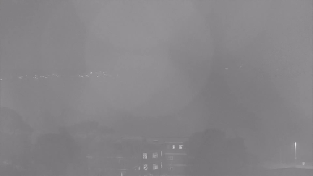A grayscale webcam image shows a foggy or misty scene. The foreground includes indistinct dark shapes, possibly trees or buildings, while a faint horizontal line of small lights suggests a distant road or urban area. The overall visibility is low, creating a soft, blurred effect throughout the image.