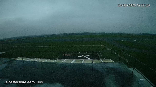 A cloudy, overcast sky covers a grassy airfield. In the foreground, there is a flat surface with a white cross marking a helicopter landing area. The background shows an expansive field with faint outlines of distant trees and possibly a road, with a few white markers scattered throughout the area. The overall lighting is dim, suggesting it may be evening. The bottom left corner displays text that reads "Leicestershire Aero Club."
