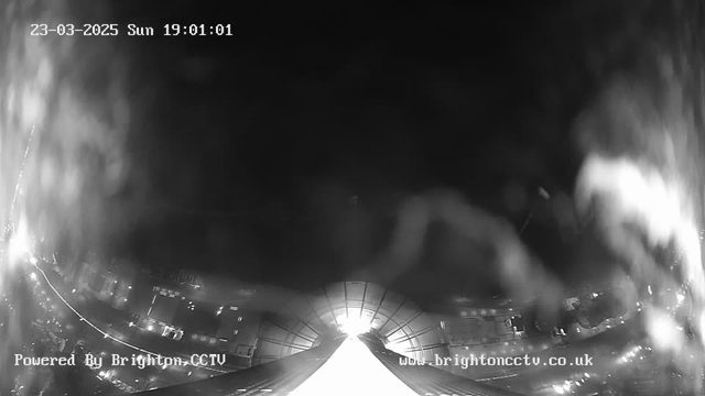 A nighttime webcam view shows a city scene illuminated by streetlights. The image is predominantly dark with areas of bright white light suggesting reflections or headlights. In the foreground, a structure with a curved roof is visible, leading towards a light source in the distance. The date and time at the top indicate it is Sunday, March 23, 2025, at 19:01:01. A watermark in the lower left corner identifies the image as powered by Brighton CCTV.