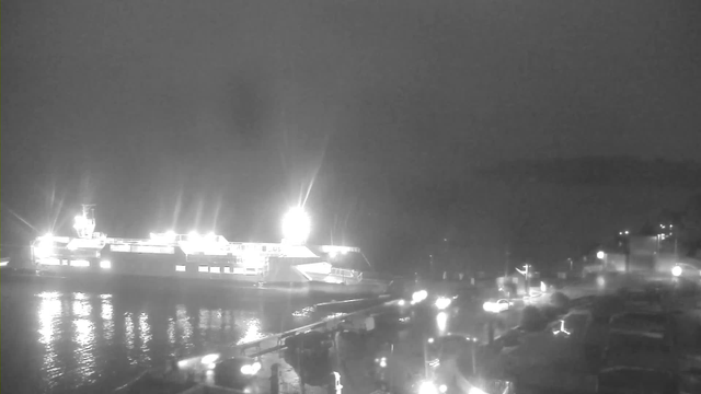 A dark, low-light scene featuring a large ship lit by bright lights. The ship is docked at a marina with reflections of the lights shimmering on the water's surface. In the foreground, there are outlines of boats and structures along the dock, and lights from nearby buildings and vehicles are faintly visible in the background. The overall atmosphere is quiet and moody, suggesting nighttime.