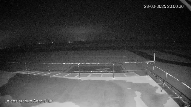 A dark image captured from a webcam at Leicester Aero Club. The scene shows a wide expanse of an airstrip, bordered by a low railing on the left side. The ground appears wet, reflecting some ambient light from distant sources in the background. The date and time displayed in the top right corner indicate March 23, 2025, at 8:00 PM. The overall ambiance is quiet and still, with limited visibility due to low light conditions.