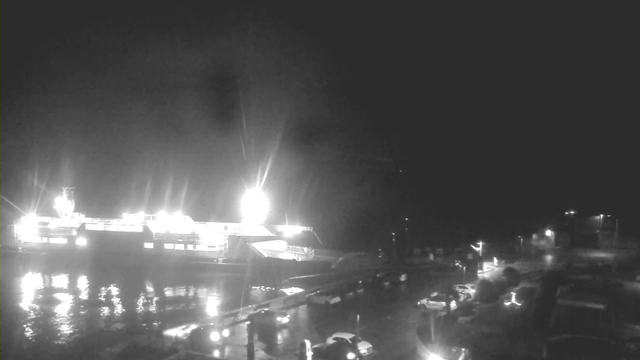 A dimly lit scene at a harbor during nighttime. A large ferry is docked, illuminated by bright lights, reflecting off the water's surface. In the foreground, several parked cars are visible along the edge of a road. The surrounding area appears quiet, with minimal background lighting and a dark sky overhead.