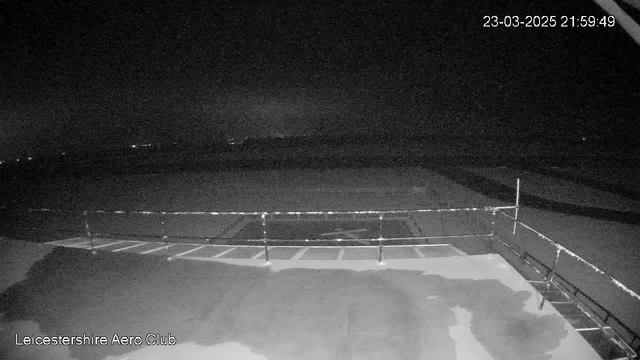 A dark scene captured from a webcam at the Leicestershire Aero Club. The image shows a flat surface, possibly an airfield or runway, with faint lines marking the ground. In the foreground, there is a railing that runs horizontally across the bottom of the image. The overall atmosphere is dim, indicating nighttime, with very little visible detail in the background. The timestamp in the corner shows the date and time as March 23, 2025, at 21:59:49.
