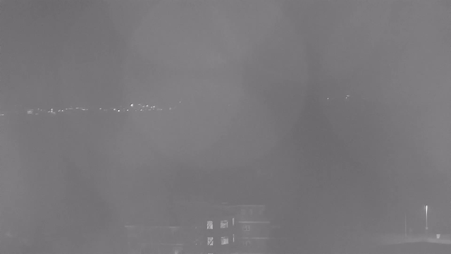 A dimly lit urban scene at night with a view of distant lights from buildings or street lights. The image appears hazy, with a blurred effect dominating the foreground and faint outlines of structures and trees. There are several small, bright spots in the distance, suggesting the presence of light sources.