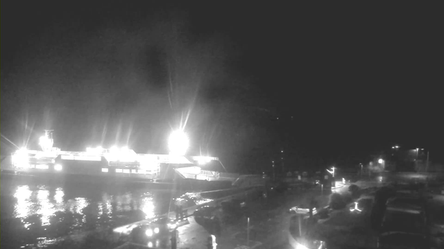 A nighttime view of a harbor area with a large, illuminated ferry docked at the pier. The ferry is bright, with strong beams of light shining from it, reflecting off the dark water. In the foreground, there are several vehicles parked along the wet pavement. The overall scene is dark with limited visibility, highlighting the contrast between the lights of the ferry and the surrounding shadows.