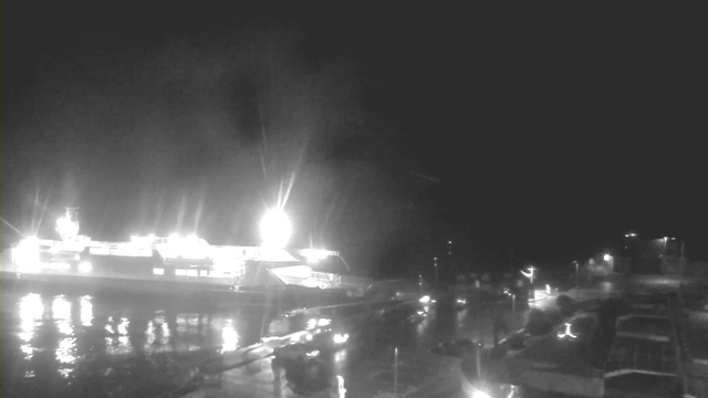 The image shows a nighttime scene at a port. A large ferry or cargo ship is docked on the left, illuminated by bright lights. Reflections of these lights shimmer on the water. The right side of the image features a harbor area with several smaller vessels, and there are additional lights along the shore. The overall atmosphere is dark, indicating nighttime.