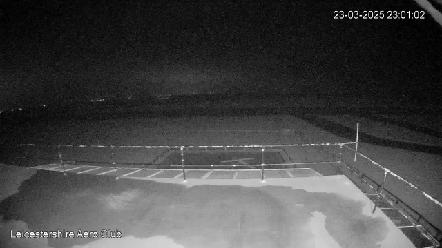 A dark scene captured by a webcam at night, showing a flat surface that appears to be an airfield or runway. A metal railing is visible in the foreground, with one section open. In the background, there are indistinct shapes that may represent distant hills or buildings, with faint lights from some unknown sources. The image lacks color and detail due to low light conditions. The bottom left corner has text indicating "Leicestershire Aero Club." The timestamp indicates the image was taken on March 23, 2025, at 23:01:02.