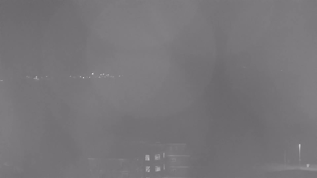 A dimly lit night scene captured from a webcam. In the foreground, a building is visible with a few lighted windows. In the background, a line of distant lights suggests a shoreline or road, but details are obscured and hazy, creating a foggy atmosphere.