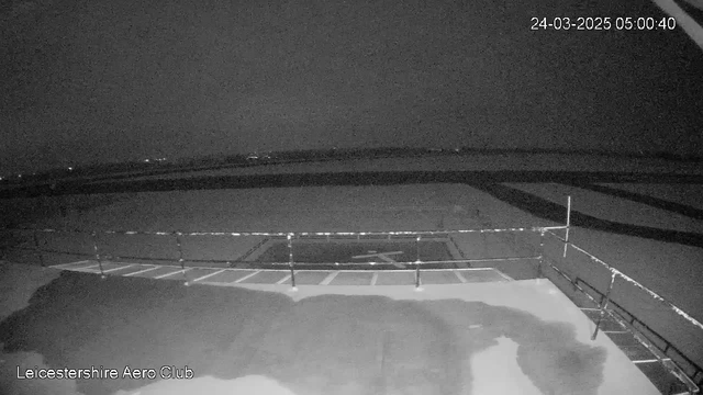 A dimly lit outdoor scene captured from a webcam at the Leicestershire Aero Club, showing a flat surface with a large, partially illuminated area in the foreground. There is a railing running along the edge of the surface, and faint outlines of shadows and distant lights can be seen in the background. The time displayed in the top right corner indicates it is early morning on March 24, 2025.