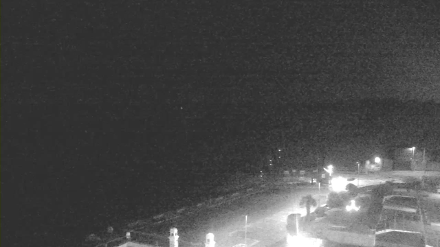 A dark scene captured by a webcam at night, showing a mostly empty street by the shoreline. There are faint lights indicating buildings and street lamps, but overall, the image is predominantly black with little visible detail. The water is not discernible, blending into the darkness.