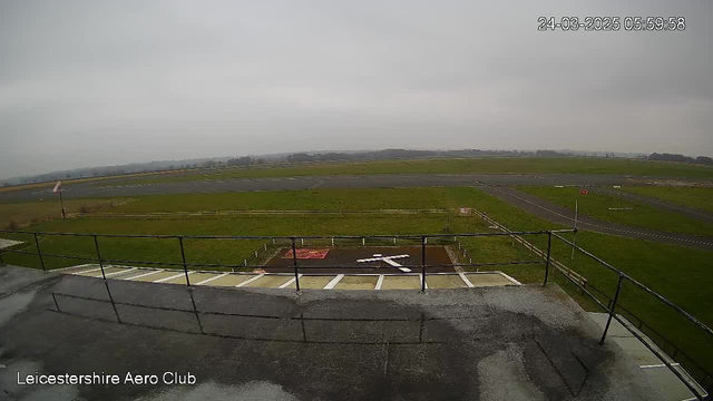 A view from a webcam overlooking a grassy airfield on a cloudy day. The foreground shows a low wall and a railing. The airfield features a cross-shaped marking on the ground, with a red square and a small sign nearby. In the background, the airfield is bordered by a wooden fence and a runway is visible, along with a windsock indicating wind direction. The sky is overcast, contributing to a muted light atmosphere. The timestamp in the upper right corner shows the date and time.