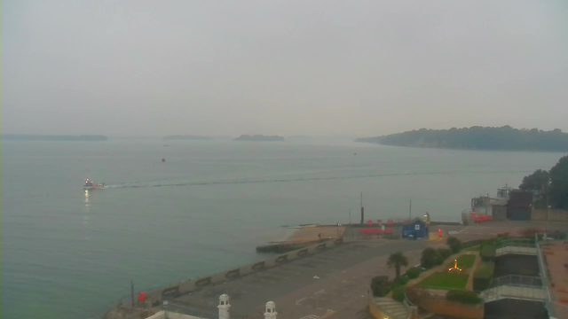 A cloudy scene over a calm body of water, with several small islands visible in the distance. In the foreground, there is a concrete walkway and a dock. A small boat is traveling across the water, leaving a gentle wake behind it. To the right, there are some structures, including a blue building and orange equipment, along with a landscaped area featuring low hedges and plants. The overall atmosphere appears serene and slightly overcast.