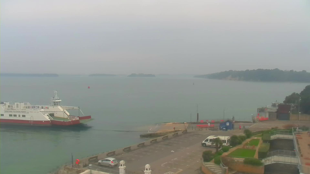 A view of a calm body of water with a foggy sky above. In the foreground, a large ferry boat is docked at a pier with a red and white color scheme. The pier has vehicles parked and several people visible near the water's edge. Beyond the pier, small islands can be seen in the distance, partially enveloped by mist. The shoreline features greenery and well-maintained gardens.