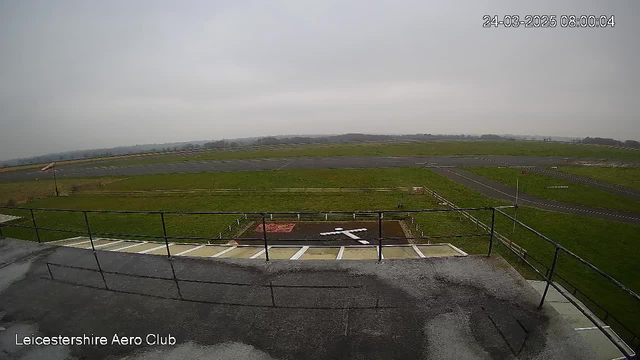 A view from a webcam overlooking an airfield. The scene shows a large expanse of grassy land and a portion of a runway. In the foreground, there is a flat, concrete area with white lines indicating a helicopter landing zone, marked with a large white cross. Beyond the airfield, a faint horizon can be seen, shrouded in a light mist or overcast sky. The surrounding area features some implements of fencing. The overall atmosphere appears calm and overcast, indicative of early morning light.