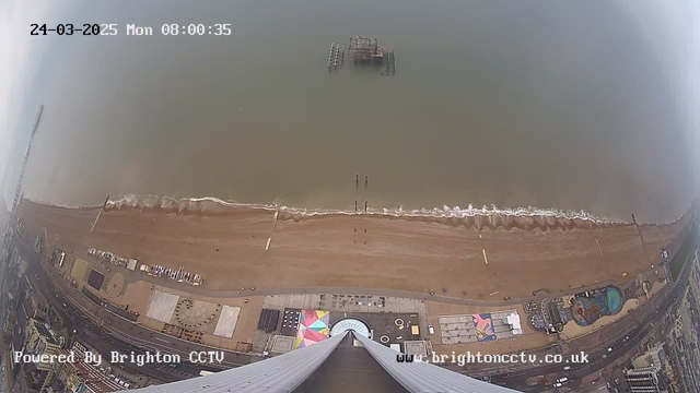 A high-angle view of a beach and the ocean taken from a webcam. The sandy beach stretches horizontally across the bottom third of the image, with a few dark vertical posts emerging from the water. Gentle waves are lapping against the shore. Visible on the beach are colorful structures and possibly people walking. The water appears murky and light brown, with hazy skies above. In the distance, a damaged pier is partially submerged. The image is timestamped with the date and time at the top.