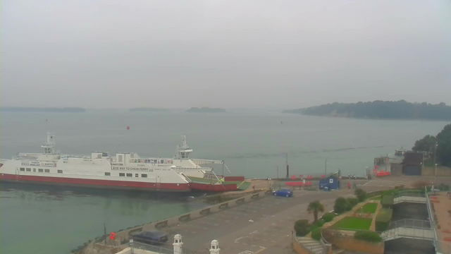 A foggy shoreline scene featuring a white and red ferry docked at a harbor. The ferry is large and has a ramp extending towards the dock. In front of the ferry, there is a paved parking area with a blue car parked. Along the waterfront, there are a few palm trees and grassy areas. The water appears calm and extends to the horizon, where faint outlines of distant land can be seen. The sky is overcast and gray, contributing to the overall muted atmosphere.