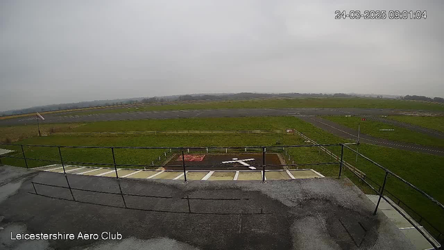 A view from the top of a building at the Leicestershire Aero Club. In the foreground, there is a fenced area with a large white cross shape marked on the ground. The background features a grassy field and a runway with a few markings. The sky is overcast, and the scene appears quiet and spacious, with no visible aircraft.