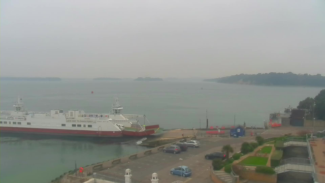 A gray, overcast scene featuring a harbor. In the foreground, a white ferry with a red bottom is docked at a pier. There is a small buoy in the water nearby. The background includes a calm sea, distant islands, and a shoreline with greenery and buildings. Several cars are parked near the ferry terminal, and a blue structure can be seen on the right side of the image. The overall atmosphere is peaceful and muted due to the clouds.
