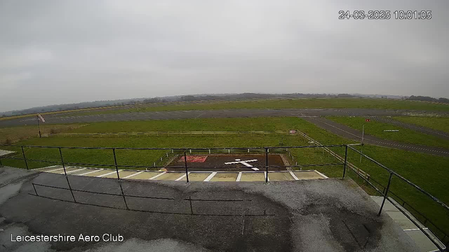 A view from a webcam situated at the Leicestershire Aero Club, capturing a cloudy sky with a grayish atmosphere. In the foreground, there is a railing, and beyond it, the airfield is visible, lined with green grass. A landing pad marked by a white cross is present on the ground, alongside a red area. The airstrip in the background is mostly asphalt and appears well-maintained, with a fence lining the perimeter. No aircraft are visible in this scene. The time displayed is 10:01 on March 24, 2025.