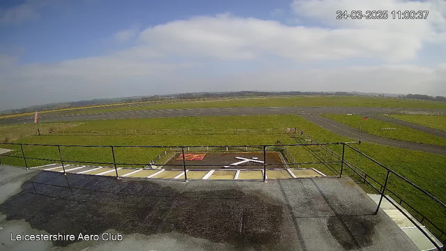 A view from a webcam at the Leicestershire Aero Club shows a large, open grassy area with a runway in the distance. The sky is partially cloudy with hints of blue. In the foreground, there are white lines marking a helipad with a large white cross. A red windsock is visible on the left side, indicating wind direction. A fence borders the grassy area, and a few signs can be seen in the distance near the runway. The overall scene is tranquil and expansive, reflecting an airfield setting.