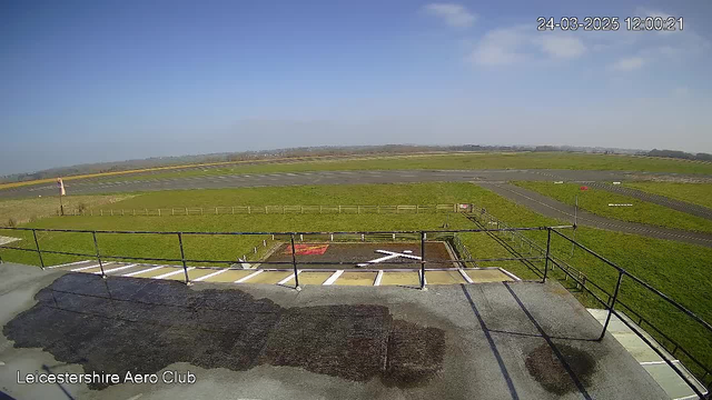 A view from a webcam at a small airport. The scene shows a large grassy area with a paved runway in the background. A helipad marked with a red “H” and a white cross is visible in the foreground. There are several small clouds in a bright blue sky. A wooden fence borders the grassy area, and there is signage indicating airport directions. The overall atmosphere is clear and sunny, with no aircraft visible in the immediate vicinity.