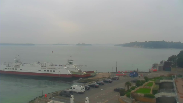 A view of a harbor under overcast skies. In the foreground, a large ferry boat is docked at the pier with a red and white color scheme. Several vehicles are parked on a nearby road, mostly dark-colored cars. The water is calm and gray, with small boats visible in the distance. On the right side, there are buildings and pathways, with some greenery along the edge. The horizon includes a wooded island.